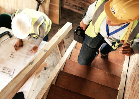 Two construction workers in hard hats and safety vests review blueprints on a wooden staircase site, one holding a cordless drill.